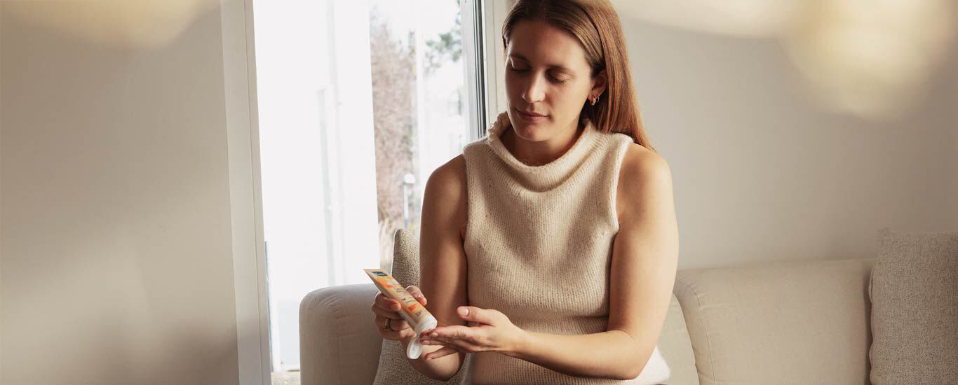 Een vrouw zit op de bank en smeert haar handen in met Kneipp-handcr&egrave;me. 
